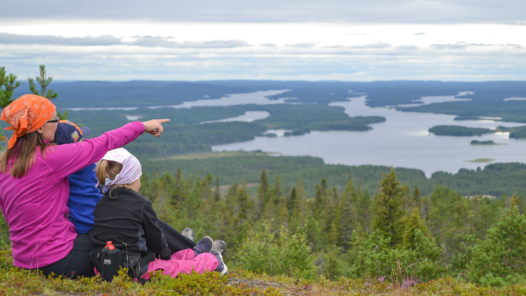 En vuxen sitter på en kulle med två barn och pekar på ett avlägset föremål med fingret.