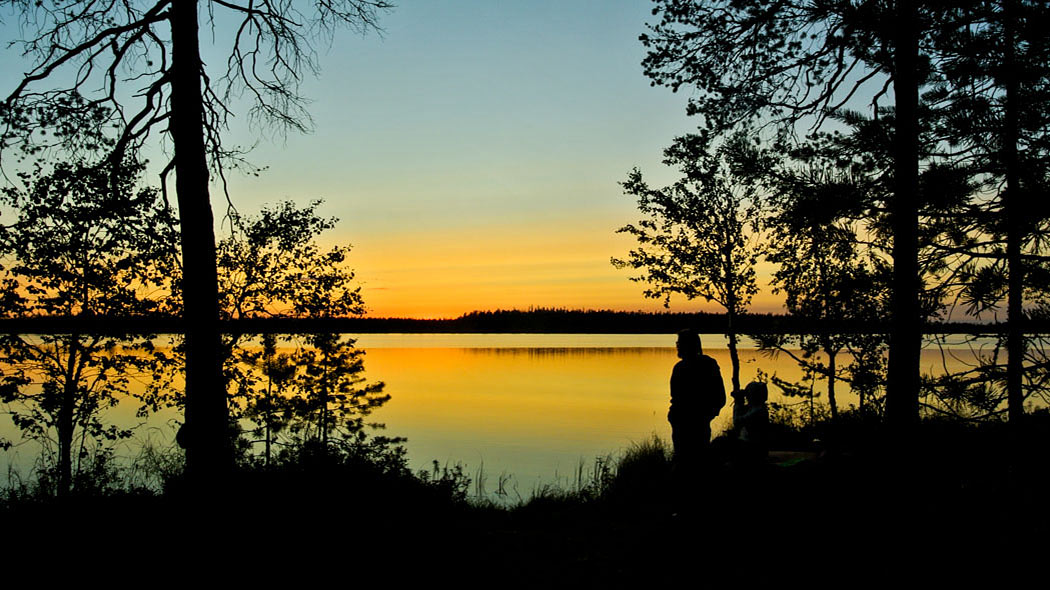 Auringonlaskussa järvimaisema ja etualalla näkyy tummana puiden sekä järvelle katselevan retkeilijän siluetti.
