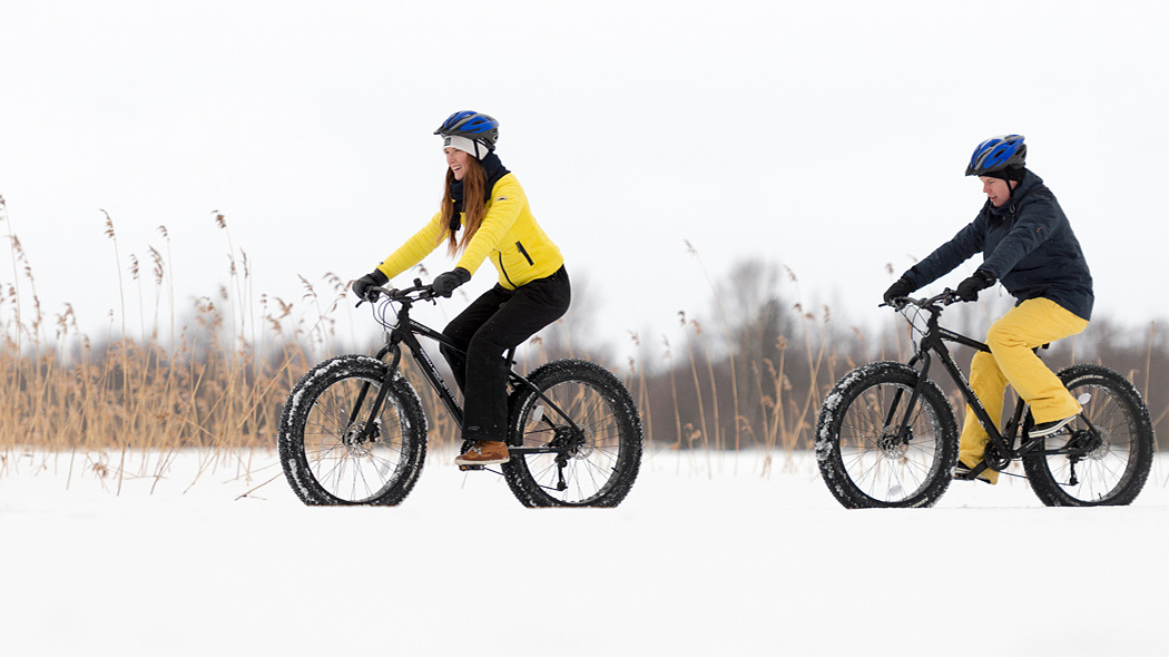 Kaksi maastopyöräilijää ajaa lumisessa maisemassa. Pyöräilijöillä on värikkäät vaatteet. Taustalla on järviruokokasvustoa.
