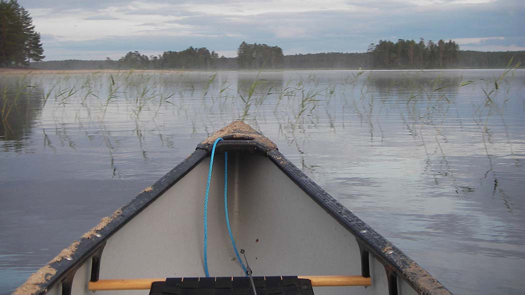 Ett dimmigt sjölandskap avbildat från en kanot. I förgrunden en kanot.