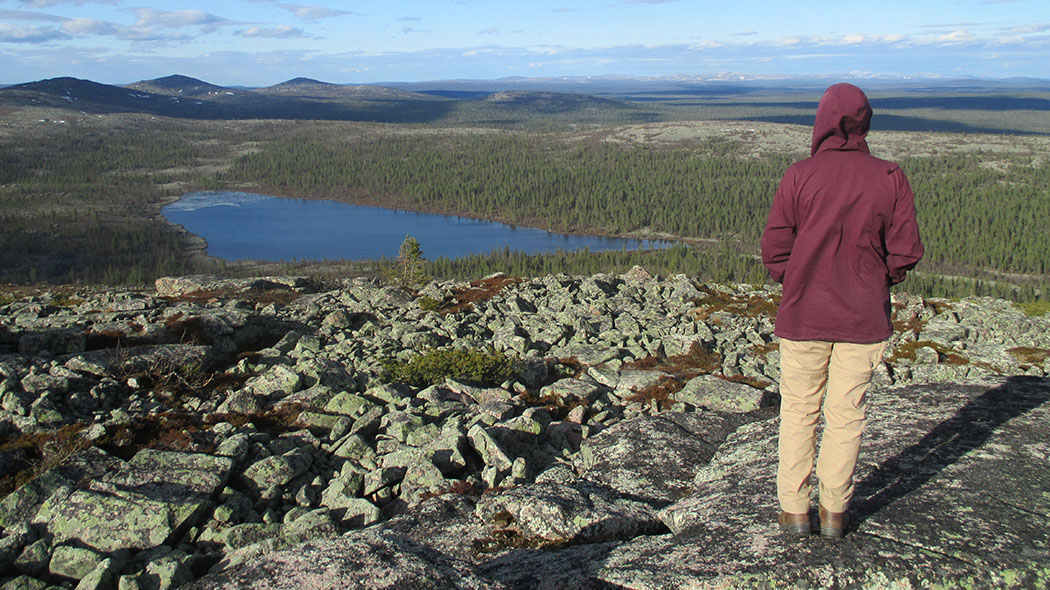 En person tittar ner från toppen av ett fjäll över landskapet som öppnar sig framför. I mitten finns en sjö och bakom den finns fjäll med spetsiga toppar.