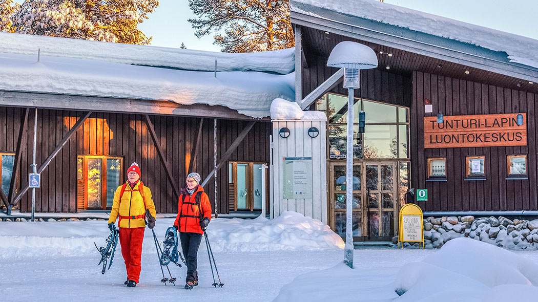 Kaksi retkeilijää kävelee talon edustalla talvella. He kantavat lumikenkiä ja sauvoja.