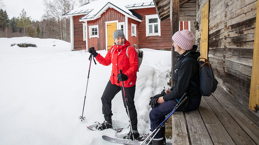 Kaksi naista talvella torpan pihalla. Toinen naisista istuu vanhan rakennuksen seinustalla olevalla penkillä ja toinen seisoo penkin vierellä lumikengät jalassaan.