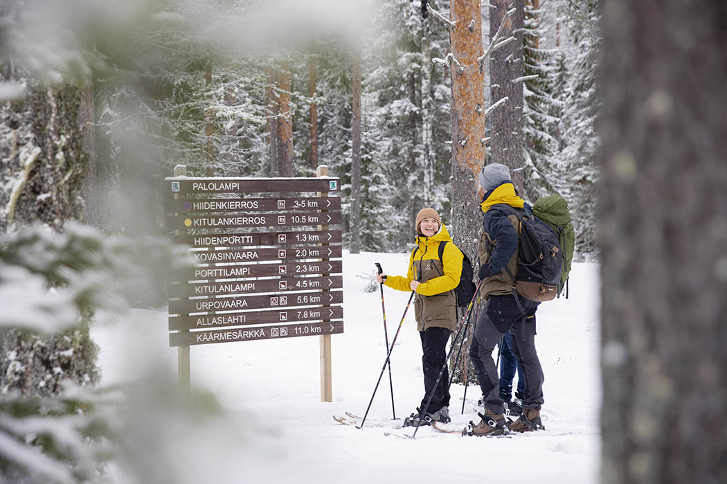 Kaksi hiihtäjää on pysähtynyt opasviitan viereen lumisessa metsässä.