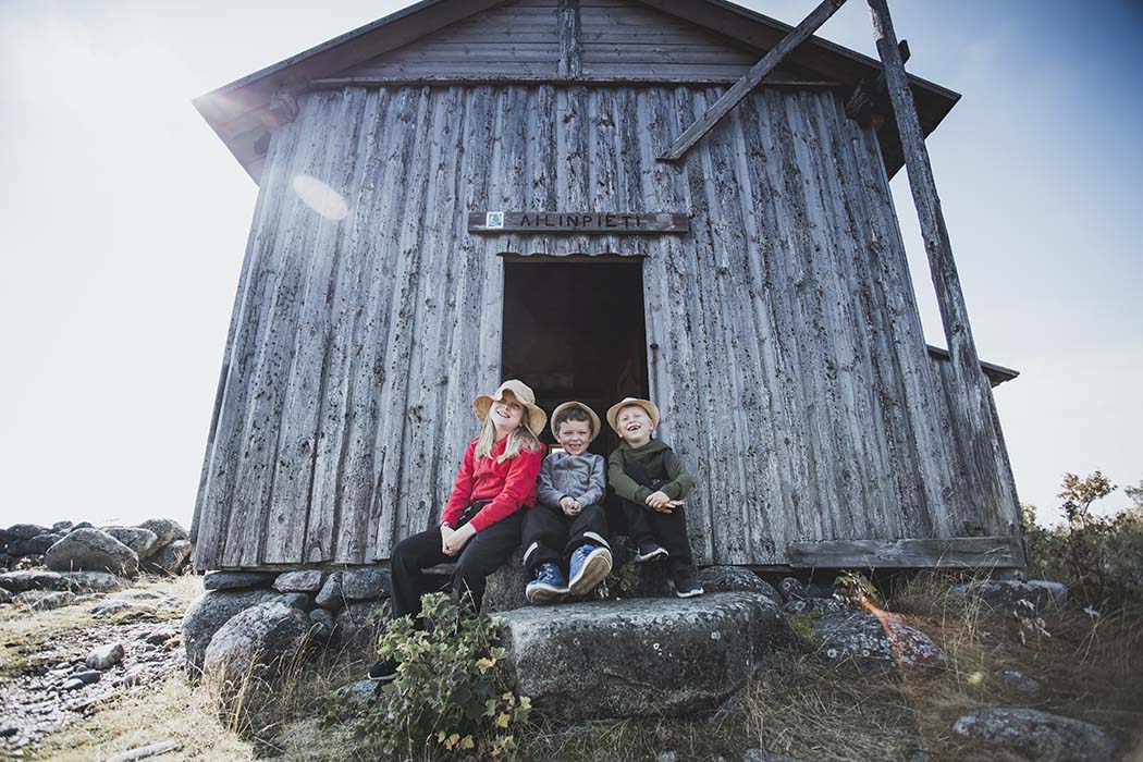 Kolme lasta istuu harmaaksi patinoituneen rakennuksen oviaukossa. Oviaukon yläpuolella on kyltti, jossa lukee Ailinpieti.