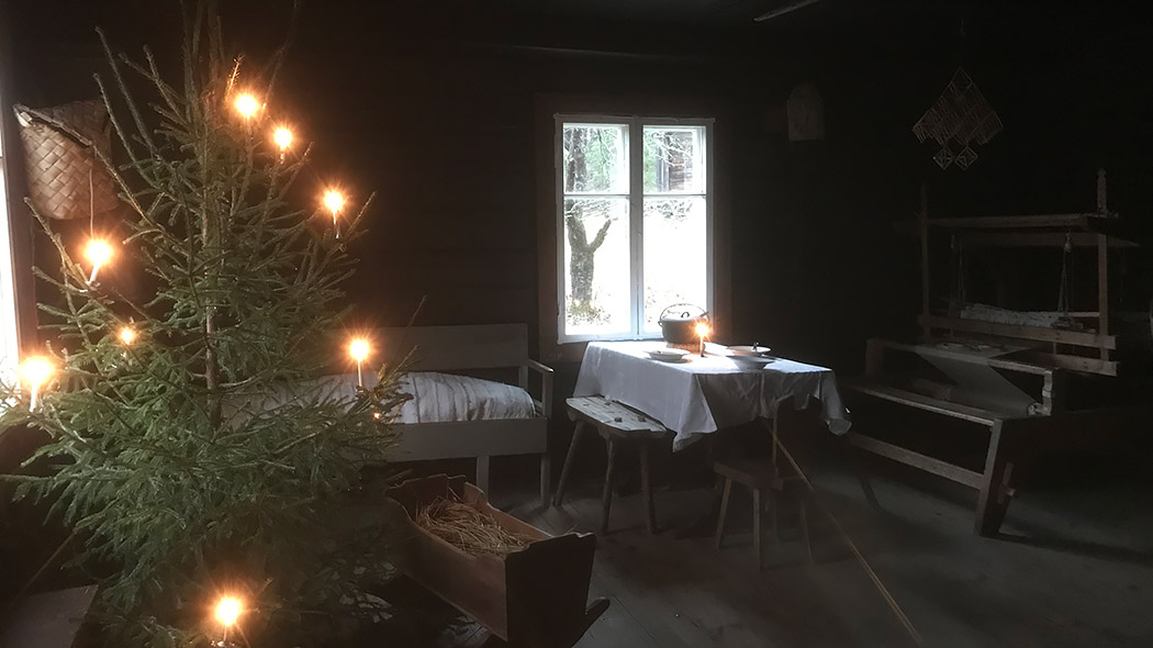 Hämärässä tuvassa on kangaspuut, pöytä, penkit, puusohva, puinen kehto sekä kynttilöin koristeltu joulukuusi.