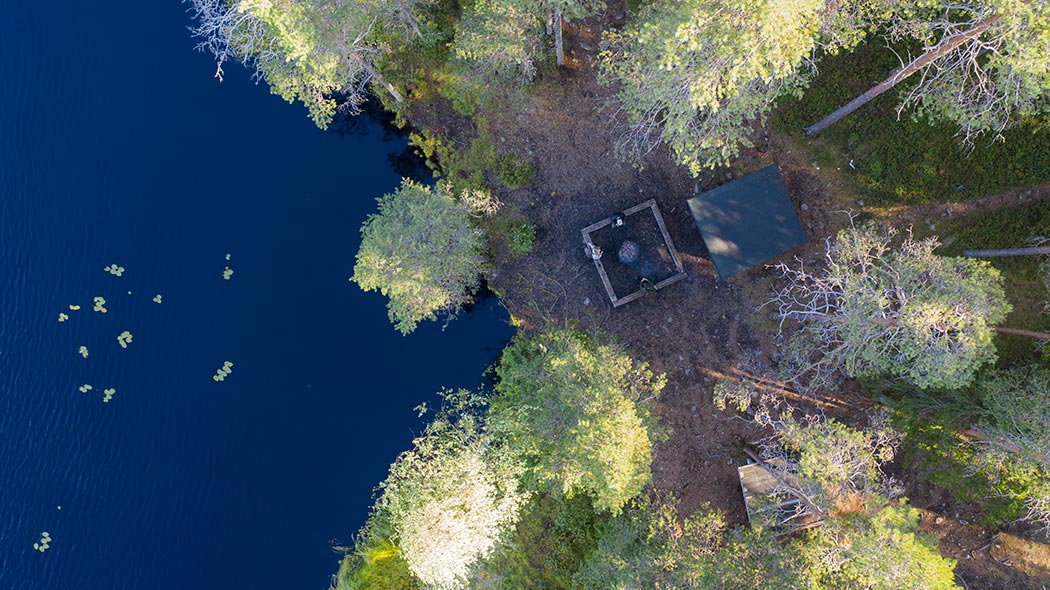 Ilmasta otetussa kuvassa järven rannassa puiden siimeksessä on laavu ja tulentekopaikka, jossa istuu kaksi ihmistä.